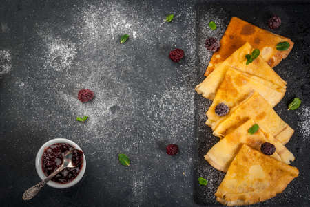 French crepes on a black slate board with fresh blackberries, mint leaves and jam, sprinkled with powdered sugar top view copy spaceの写真素材