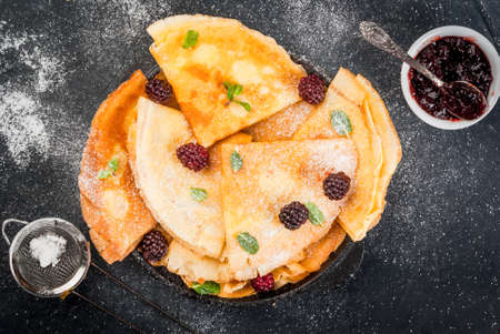 French crepes on a black rustic pan with fresh blackberries, mint leaves and jam, sprinkled with powdered sugar top view copy spaceの写真素材