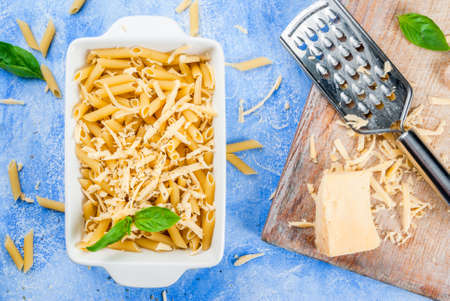 Macaroni with grated cheese in a pot ready to bake; grater and grated cheese on a cutting board, spices and tomatoes. On a light blue stone concrete table top view copy spaceの写真素材