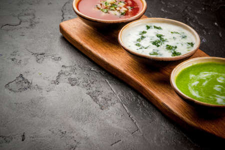 Selection of cold refreshing summer soups: tomato gazpacho, tarator, cream soup of green peas on a cutting board on a black concrete table close view copy spaceの写真素材