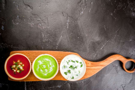 Selection of cold refreshing summer soups: tomato gazpacho, tarator, cream soup of green peas on a cutting board on a black concrete table top view copy spaceの写真素材