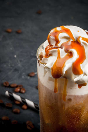 Cold coffee drink frappe (frappuccino), with whipped cream and caramel syrup, with straws and grains of coffee on a dark gray stone table, close view copy spaceの写真素材