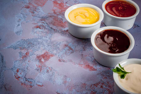 Classic set of sauces in white saucers: American yellow mustard, ketchup, barbecue sauce, mayonnaise. On grey blue with the effect of rust, concrete stone table close view, copy spaceの写真素材