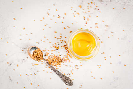 Fresh yellow American mustard in a small jar with a spoon. With mustard grains, on a white stone background, top view copy spaceの写真素材