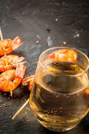 Seafood. Shellfish. Fried grilled shrimp prawns on wooden skewers, glass of white wine. Snack, appetizer. On black concrete table, close view, copy space, verticalの写真素材
