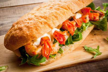Sandwich from freshly baked home-made ciabatta rolls with lettuce, greens, tomatoes, cheese sauce and grilled fried shrimps prawns. On a wooden table. Copy spaceの写真素材