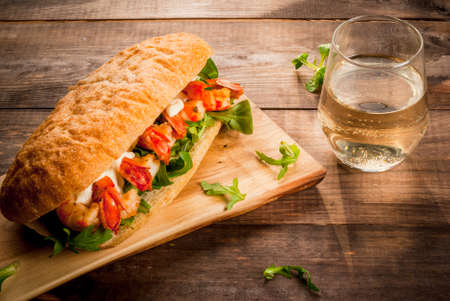 Sandwich from freshly baked home-made ciabatta rolls with lettuce, greens, tomatoes, cheese sauce and grilled fried shrimps prawns. With a glass of white wine. On a wooden table. Copy spaceの写真素材