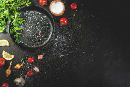 Cooking, background. Ingredients and spices for cooking dinner: tomatoes, greens (parsley), salt, pepper, onions, shallots, garlic. With a frying pan, on a black concrete table, copy space, top viewの写真素材