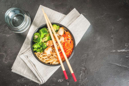 Healthy eating, diet. Buddha bowl hot salad in Asian style: noodles with soy sauce, spicy carrots, chicken, broccoli. On black concrete stone table, with chopsticks and water. Top view copy spaceの写真素材
