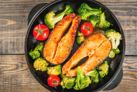 Healthy eating, diet. Baked grilled trout (salmon) with vegetable garnish - broccoli, tomatoes. In a portioned frying pan, on a wooden table. Top view copy spaceの写真素材