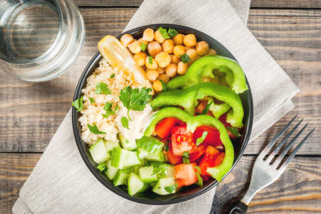 Healthy eating, diet. Vegetarian vegetable buddha bowl - couscous (quinoa), chickpeas, tomatoes, zucchini (cucumber), pepper, hummus, lemon and greens. On wooden table, with water, close top viewの写真素材