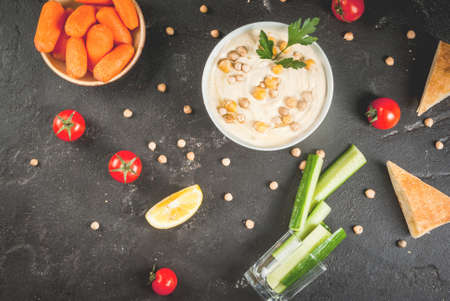 Healthy food. Vegetable sources of protein. Bowl of hummus, on black stone table, with greens, boiled and raw chickpeas. With fresh cucumber, tomatoes, carrots and bread pita. Copy space top viewの写真素材