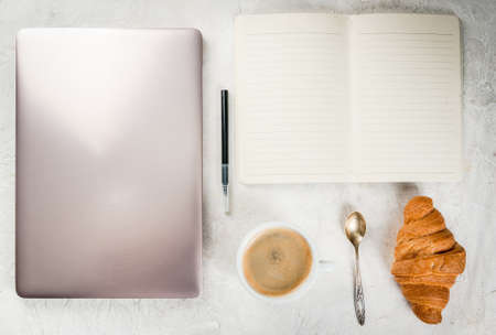 Coffee break. Breakfast or lunch at work. Desk in the office. On a white background. Laptop, notebook, pen, croissant, spoon and cup of coffee. Top view copy spaceの写真素材