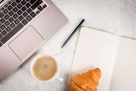Coffee break. Breakfast or lunch at work. Desk in the office. On a white background. Laptop, notebook, pen, croissant, spoon and cup of coffee. Top view copy spaceの写真素材