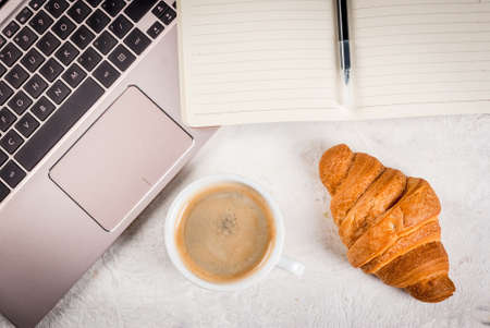 Coffee break. Breakfast or lunch at work. Desk in the office. On a white background. Laptop, notebook, pen, croissant, spoon and cup of coffee. Top view copy spaceの写真素材