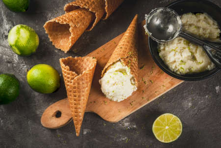 Refreshing lime sorbet in a bowl with a spoon for ice cream. With horns for ice cream (one full of ice cream), limes, grater for peel. On an old gray concrete table. Top view copy spaceの写真素材