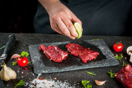 Preparation of dinner. Cooking, processing of meat (beef, tenderloin). Person (man's hands) marinates meat - sprinkles with lemon juice. Black concrete table, spices and herbs in the frame.の写真素材