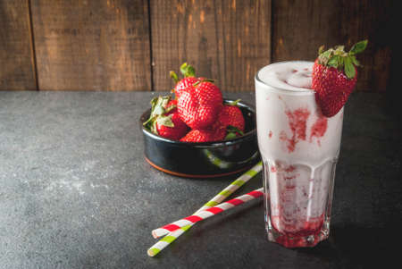 Milkshake with strawberries. In a glass on a stone black and dark wooden background. With strawberries in a bowl next to and striped bright straws. copy spaceの写真素材