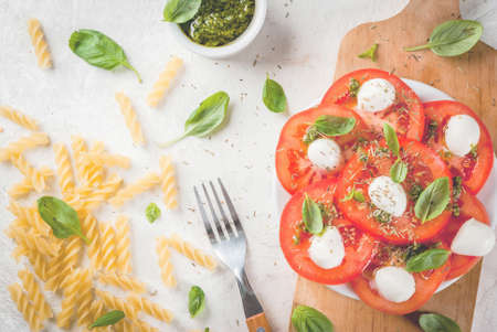 Italian food. Spring dinner for one person. Salad with caprese - tomatoes, mozzarella cheese, fresh and dried herbs, basil and pesto sauce. With fork. On a white stone table.の写真素材