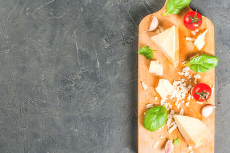 Ingredients of Italian cuisine.Parmesan grated and a piece, with a grater, basil leaves, garlic and cherry tomatoes on cutting board on dark concrete table. Top view copy spaceの写真素材