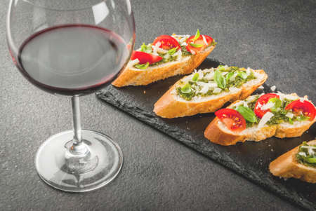 Italian food. Snacks. Antipasto. Bruschetta with pesto sauce, parmesan cheese, tomatoes and fresh basil. On black table, on slate board. With glass of red wine. Copy space selective focusの写真素材