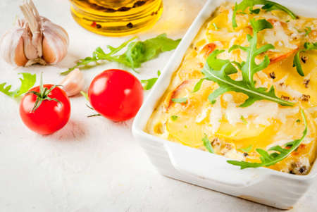 Casserole. French cuisine. Homemade potato gratin in a ceramic frying pan for baking. On a white marble stone background. With leaves of fresh arugula, tomatoes, olive oil. Copy spaceの写真素材