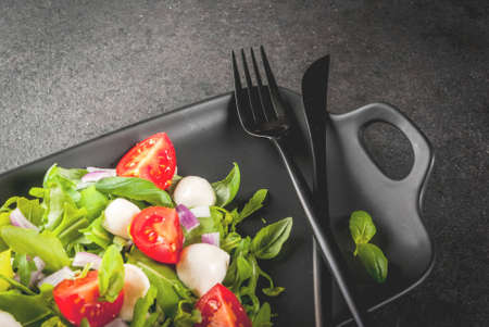 Light spring salad from leafy vegetables, with arugula, basil, mozzarella cheese balls and tomatoes. On a black rectangular plate, with a knife and fork. On a black stone table. Top view copy spaceの写真素材