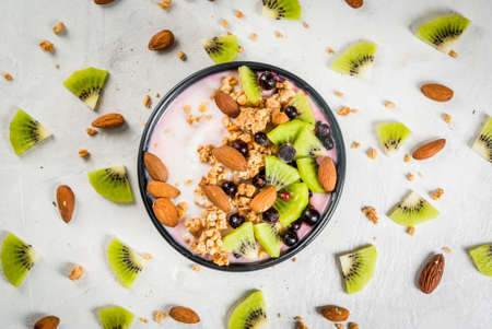 Healthy diet breakfast: smoothies bowl, with yogurt, fresh blueberries (black currant), kiwi, granola oatmeal, almond nuts. On a white stone table, with a glass of water and ingredients. Top viewの写真素材