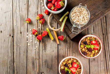 Healthy breakfast. Oatmeal granola crumble with rhubarb, fresh strawberries and blackberries, seeds and ice cream in baked bowls, with mint, on a wooden rustic table in old tray, top view copy spaceの写真素材