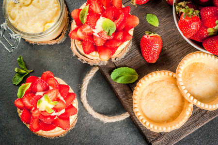 Summer and spring dessert. Home pies tartlets with custard and strawberries, decorated with mint and powdered sugar. On black stone table, rustic, with wooden board, tray. Copy space top viewの写真素材