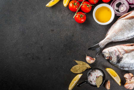 Fresh raw organic fish Dorado, sea bream with ingredients for cooking lemon, tomatoes, onion, salt, pepper, bay leaf, greens, oil. On black stone table. Top view copy spaceの写真素材