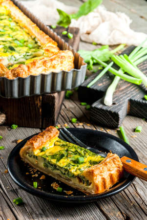 French home cooking. Casserole. Pie. Quiche lorraine from puff pastry, with young green onions and spinach. On the old wooden rustic table. Cut. In form for baking. Plate, fork, knife. Copy spaceの写真素材