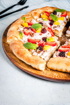 Summer snacks. Food for party. Fruit pizza with cream, currants, yogurt, strawberries, mango, peaches, bananas, blackberries, chocolate, walnuts, mint. On light blue table. Copy spaceの写真素材