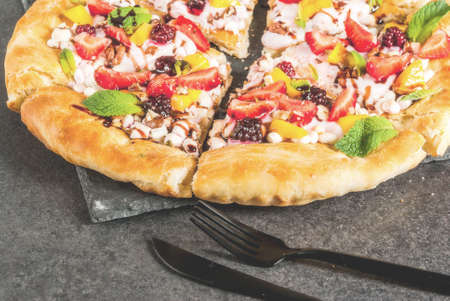 Summer snacks. Food for party. Fruit pizza with cream, currants, yogurt, strawberries, mango, peaches, bananas, blackberries, chocolate, walnuts, mint. On black table. Copy spaceの写真素材