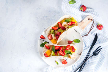 Summer snacks. Food for a party. Fruit tacos with strawberries, mangoes, bananas, chocolate, mint. On a light blue concrete table. Copy spaceの写真素材