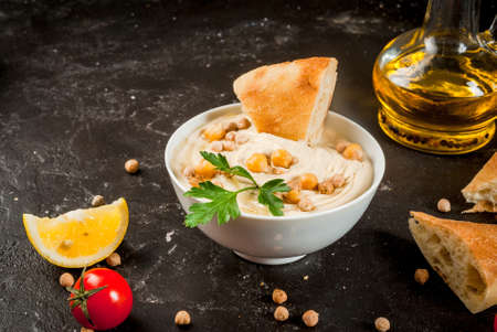 Healthy food. Vegetable sources of protein. Bowl of hummus, on black stone table, with greens, boiled and raw chickpeas. With fresh cucumber, tomatoes, carrots and bread pita. Copy spaceの写真素材