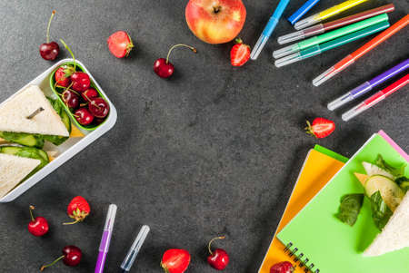 Back to school. A hearty healthy school lunch in a box: sandwiches with vegetables and cheese, berries and fruits (apples) with notebooks, colored pens on a black table. Copy space top viewの写真素材