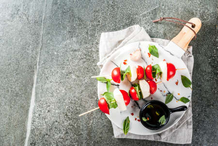 Italian dinner. Homemade salad caprese - tomatoes, fresh basil, mozzarella cheese, olive oil. On a black stone table. Served on skewers. On a marble plate. Copy space top viewの写真素材