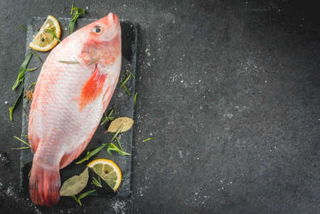Raw fish red tilapia on a chopping board on a black stone table, with spices, lemon and herbs for...