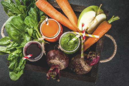 Vegan diet food. Detox drinks. Freshly squeezed juices and smoothies from vegetables: beets, carrots, spinach, cucumber, apple. On dark stone background, wooden tray, ingredients. Copy space top viewの写真素材