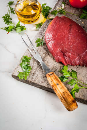 Cooking beef fillet tenderloin. Large whole piece of veal fillet on an old cutting board with a fork for meat, spices (salt, pepper, parsley, garlic, onions, tomatoes). Copy spaceの写真素材