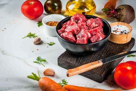 Meat, beef. Fresh raw chopped goulash, beef cubes in a bowl. Spices (salt, pepper), tomatoes, garlic, onions. On a white marble table, with a fork for meat and a knife. Copy spaceの写真素材