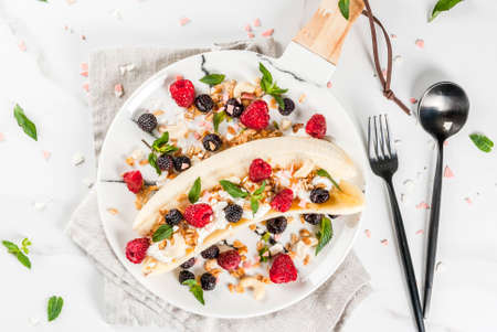 Summer fruit berry breakfast. Healthy banana split breakfast with cream cheese, raspberries, blackberries, mint, white and pink chocolate. On white marble plate, marble table. Top view copy spaceの写真素材