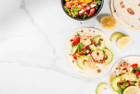 Seafood. Mexican food. Tortilla tacos with traditional homemade salsa salad, parsley, fresh lemon, avocado and grilled shrimp pawns. On a white marble background. Top view copy spaceの写真素材