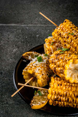 Summer food. Ideas for barbecue and grill parties. Grilled corn grilled on fire. With a sprinkle of cheese (mexican elotes), hot chili pepper, lemon. On a dark stone table, black plate Copy spaceの写真素材