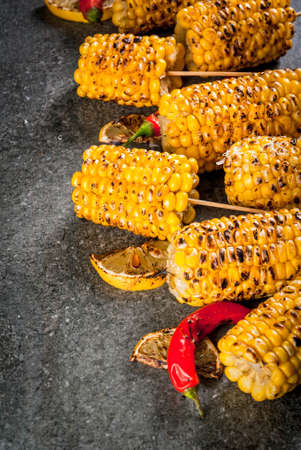 Summer food. Ideas for barbecue and grill parties. Grilled corn grilled on fire. With a sprinkle of cheese (mexican elotes), hot chili pepper and lemon. On a dark stone table. Copy spaceの写真素材
