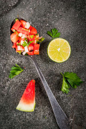 Mexican food. Summer berries and fruits. Vegetable salsa salad with watermelon, tomatoes, onions, greens, lime. In a spoon on a black stone table, with the ingredients. Top view copy spaceの写真素材