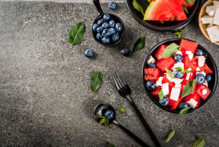 Summer fruit berry salad: watermelon, blueberries, mint, feta cheese and balsamic sauce. On a black bowl, black stone table. With ingredients, fork, knife. Copy space top viewの写真素材