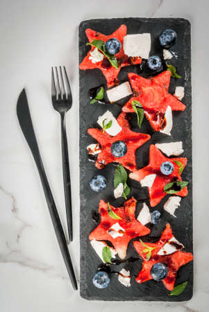 Summer fruit berry salad: watermelon sliced in the form of stars, blueberries, feta cheese and balsamic sauce. On a black slate, on a white marble table. With a fork and knife. Top view copy spaceの写真素材