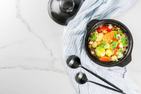 Healthy eating. Dietary vegan dinner. Homemade soup with fresh spring and summer vegetables cabbage, broccoli, green peas, carrots, celery. In black serving pan, white marble table. Copy spaceの写真素材